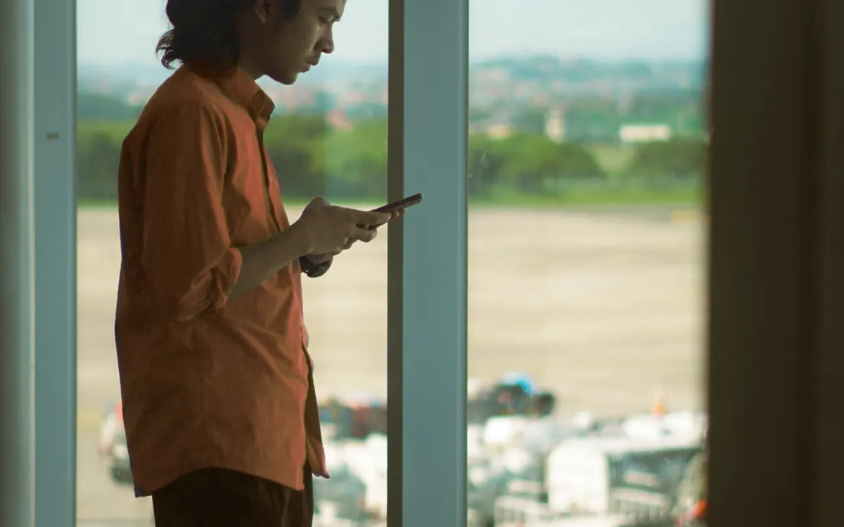 Homem em pé perto da janela do aeroporto usando o celular