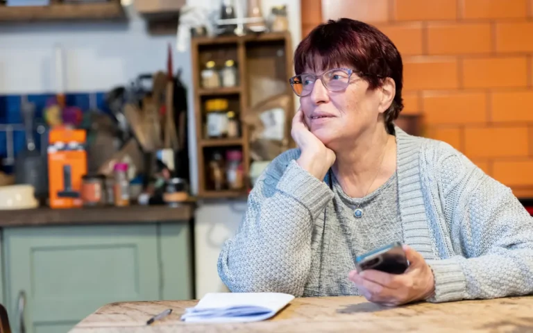 Senhora sentada à mesa da cozinha segurando o celular, pensativa